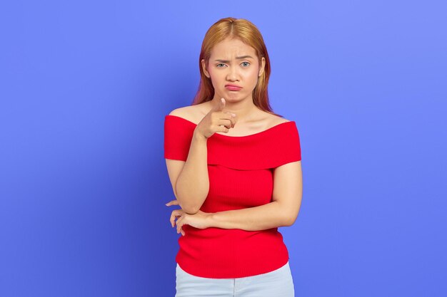 Portrait of confused young Asian woman with short blonde hair in red dress standing and pointing at camera with finger isolated over purple background