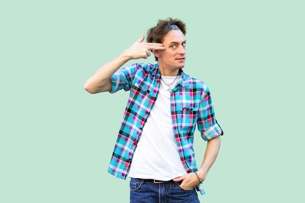Portrait of confused young man in casual blue checkered shirt headband standing with pistol gun gesture on head and looking with failure face. indoor studio shot, isolated on light green background.