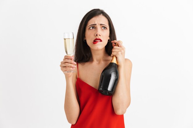 Portrait of a confused young woman in dress holding glass and bottle of champagne isolated