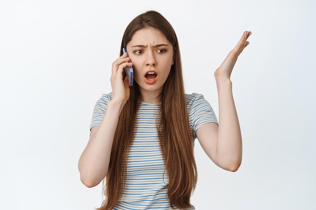 Portrait of confused young woman talking on mobile phone, raise hands in frustration, argument on smartphone, white background