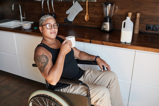 Portrait of contemporary tattooed woman in wheelchair enjoying coffee at home and looking at camera, copy space