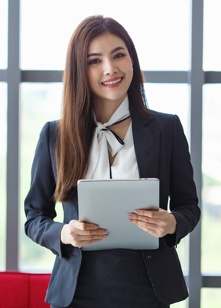 Portrait of content Asian female entrepreneur or secretary in elegant suit standing with positive friendly smile holding netbook in hall of business center and looking at camera.