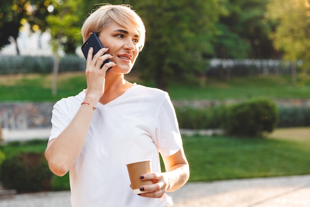 Portrait of content attractive woman wearing casual clothing smiling, while talking on mobile phone and holding takeaway coffee during walk in green park