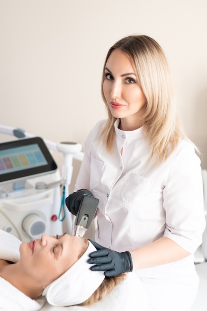 Portrait of content blond-haired beautician doing skin treatment procedure to mature woman with facial machine