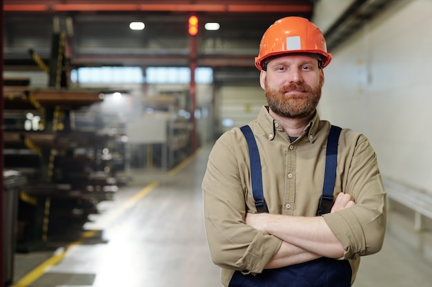 Portrait of content brutal bearded factory man in orange hardhat standing with crossed arms in workshop