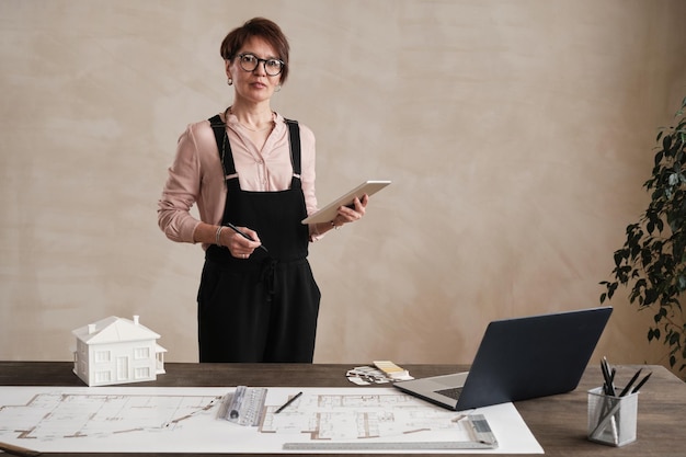 Portrait of content Caucasian interior designer in bodysuit standing with tablet and thinking of new project implementation