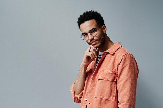 Photo portrait of content fashionable young black man in casual outfit and ring touching chin against gray