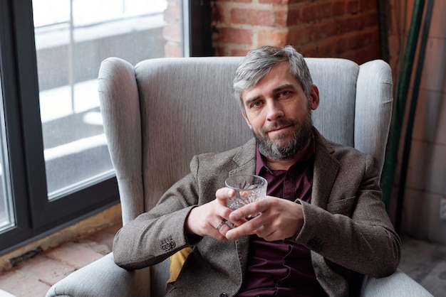 Portrait of content handsome bearded man with gray hair sitting armchair and holding whisky glass