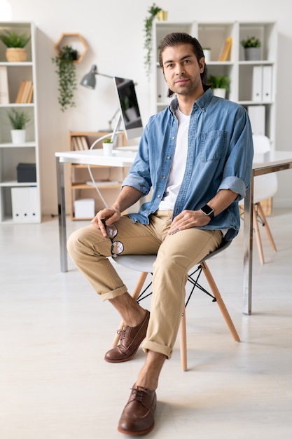 Портрет довольного красивого молодого брюнет в джинсовой рубашке, сидящего на стуле и держащего очки в современном офисе