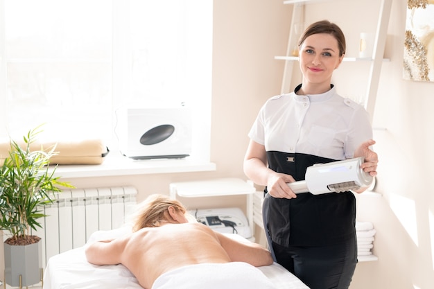 Portrait of content LPG specialist with machine handle standing at client lying on massage table in cozy room