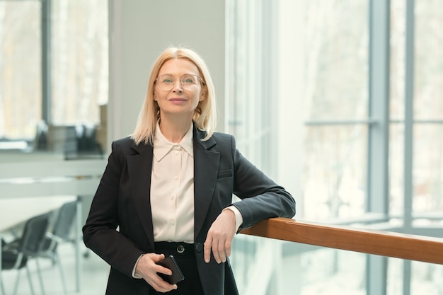 Portrait of content mature blond-haired business lady holding smartphone in modern office