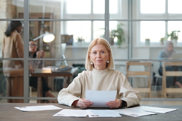 Портрет довольного зрелого менеджера по персоналу со светлыми волосами, сидящего за столом и читающего резюме кандидатов в современном офисе open space