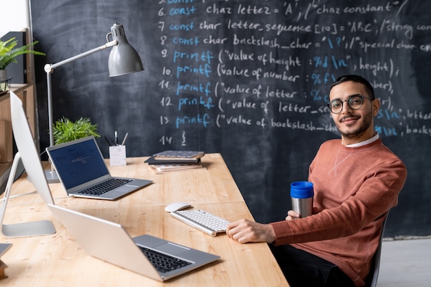 Portrait of content young Arabian coder with beard sitting with thermos cup at table with modern computers in own office