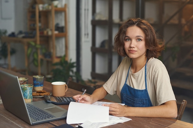 Portrait of content young caucasian pottery workshop owner in apron working with invoices using calc...