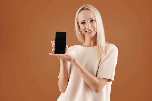 Portrait of content young Caucasian woman with blond hair standing against brown background and presenting new smartphone app