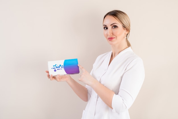 Portrait of content young female cosmetologist in lab coat recommending filler product against isolated background