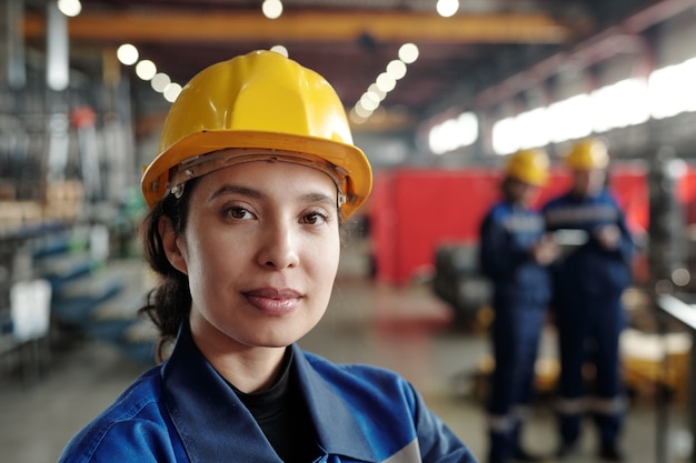 工場で働く黄色いヘルメットの若い混血女性のコンテンツの肖像画、彼女は大きな工業店に立っています