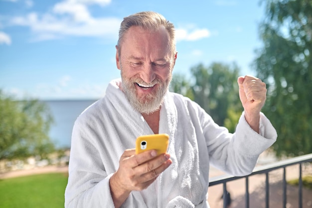 Ritratto di un gentiluomo soddisfatto in accappatoio appoggiato alla ringhiera del balcone guardando il suo smartphone