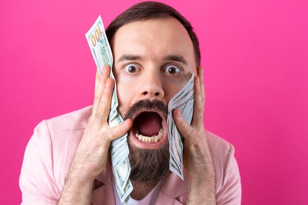 Portrait of a contented young businessman with a beard dressed in a pink jacket showing us dollar banknotes against a red studio background Taste smell of money