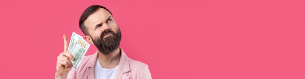 Foto ritratto di un giovane uomo d'affari soddisfatto con la barba vestito con una giacca rosa che ci mostra le banconote in dollari su uno sfondo rosso dello studio gusto odore di denaro