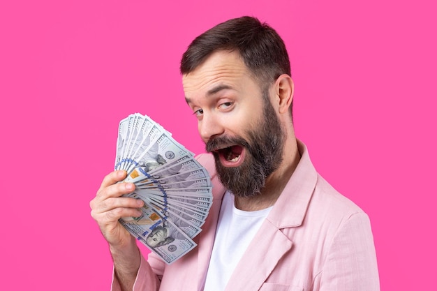 赤いスタジオの背景にドル紙幣を見せてくれるピンクのジャケットに身を包んだあごひげを生やした満足のいく青年の肖像画お金の匂いを味わう