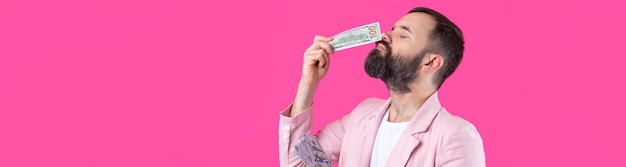 Portrait of a contented young businessman with a beard dressed in a pink jacket showing us dollar banknotes against a red studio background taste smell of money