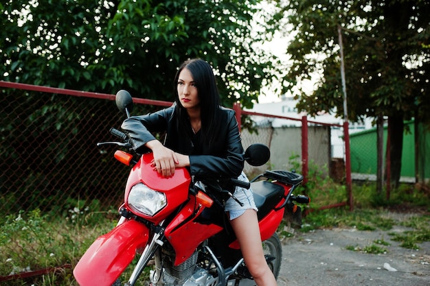 Portrait of a cool and awesome woman in dress and black leather jacket sitting on a cool red motorbike