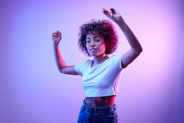 portrait of cool kazakh girl with curls and tattoo dancing and posing in neon light isolated on studio background