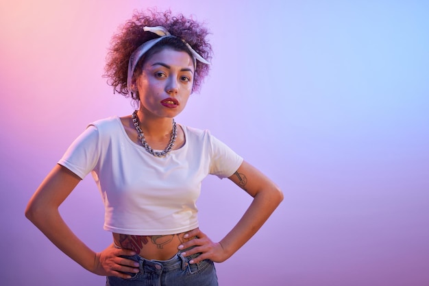 Portrait of cool Kazakh girl with curls and tattoo posing in neon light isolated on studio background