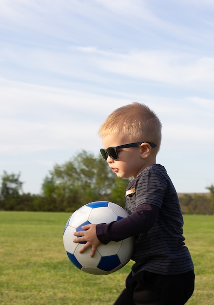 Portrait of cool toddler child in sunglasses standing in grass field stepping one leg on soccer ball. Stylish little football player at playground or park. Active childhood and sport for kids concept