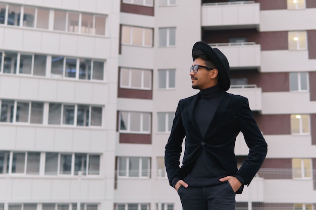 Portrait of a cool young man in black suit and hat walking on the background building