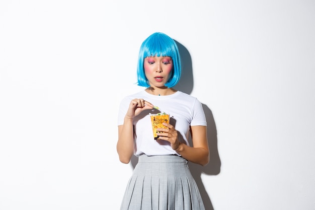 Портрет кокетливой азиатской девушки в синем парике, празднующей хеллоуин, ест сладости из кошелька или кошелька, стоя.