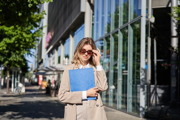 Portrait of corporate woman in sunglasses and beige suit\
holding blue folder with office documents g