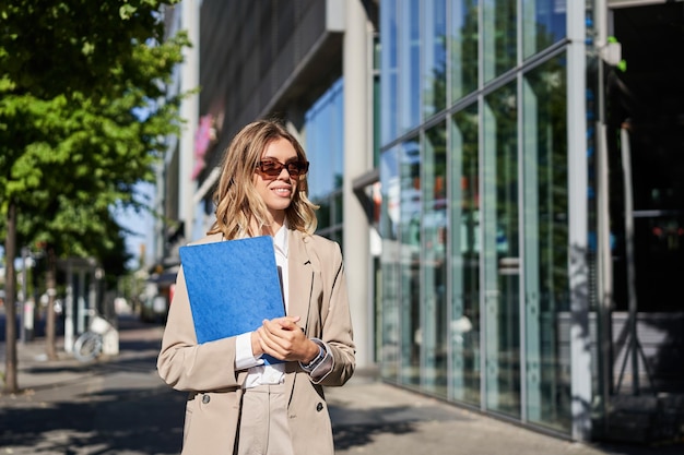 Portrait of corporate woman in sunglasses and beige suit holding blue folder with office documents g