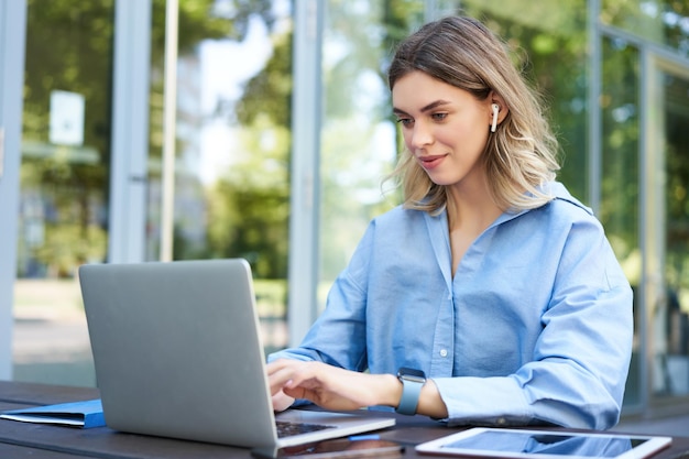 Portrait of corporate woman working outside office on remote\
sitting with laptop wireless earphones