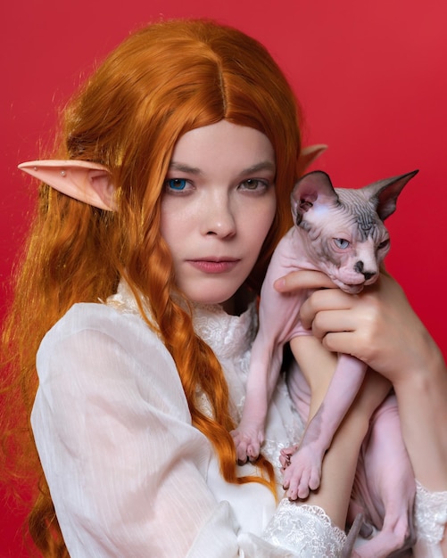 Portrait of cosplayer elf young female looking at camera thoughtfully holding Sphinx cat in hands