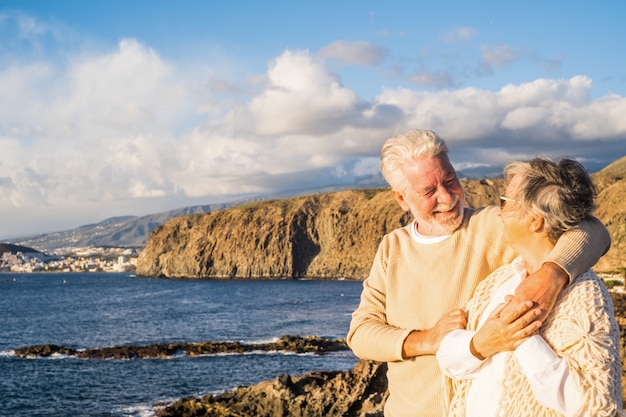Portrait of couple of mature and old people enjoying summer at the beach looking to the sea smiling and having fun together with the sunset at the background. Two active seniors traveling outdoors.
