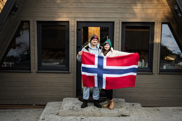 Portrait of couple outside cabin house holding Norway flag Scandinavian culture norwegian people