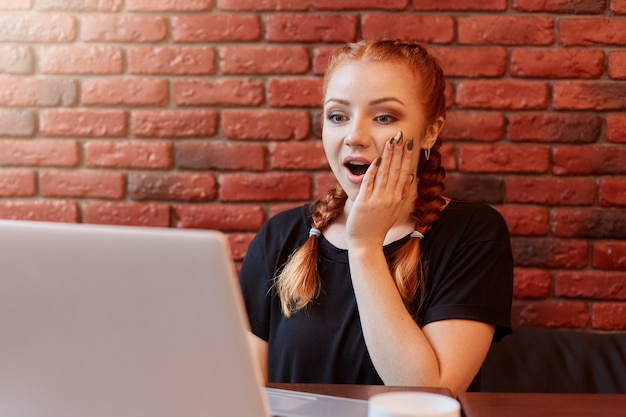 Portrait of crazy beautiful red haired young woman sitting and\
looking at her laptop display with opened mouth and big eyes with\
brick wall on space