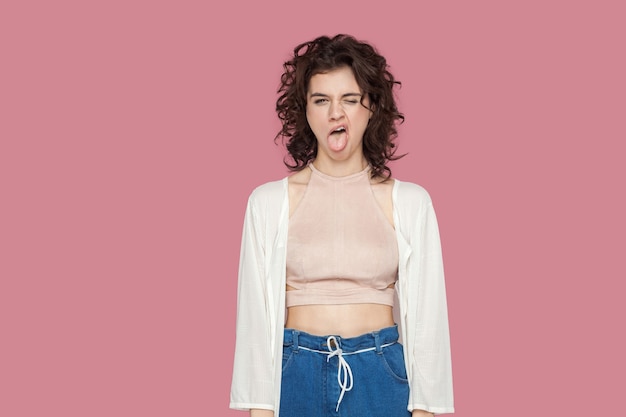 Photo portrait of crazy funny beautiful brunette young woman with curly hairstyle in casual style standing, tongue out and looking with foolish face. indoor studio shot isolated on pink background.