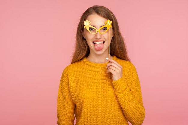 Портрет сумасшедшей смешной радостной рыжей девушки в повседневном свитере в поддельных бумажных очках, с высунутым языком, корчащим рожи и весело проводящей маскарадную студию, снятую на розовом фоне