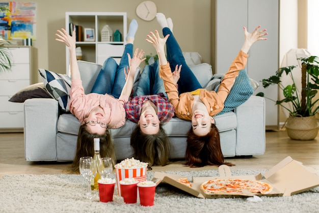 Портрет шальных смешных 3 женщин в вскользь обмундировании лежа вверх ногами на софе держа руки вверх и смотря камеру крытую. Домашняя вечеринка с пиццей