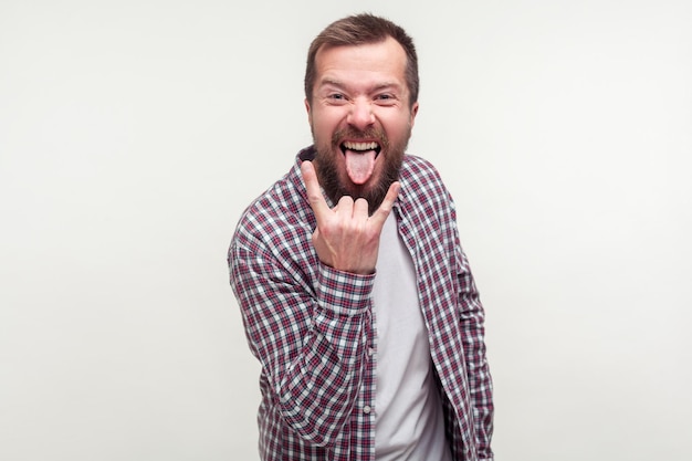 Portrait of crazy optimistic bearded man in plaid shirt showing rock and roll sign hand gesture and sticking out tongue, feeling to be cool rocker. indoor studio shot isolated on white background