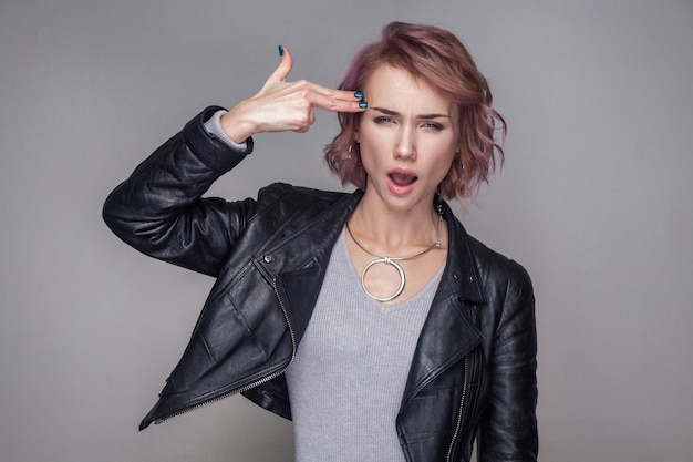 Portrait of crazy woman with short hairstyle and makeup in casual style black leather jacket standing and looking at camera with pistol gun gesture. indoor studio shot, isolated on grey background.