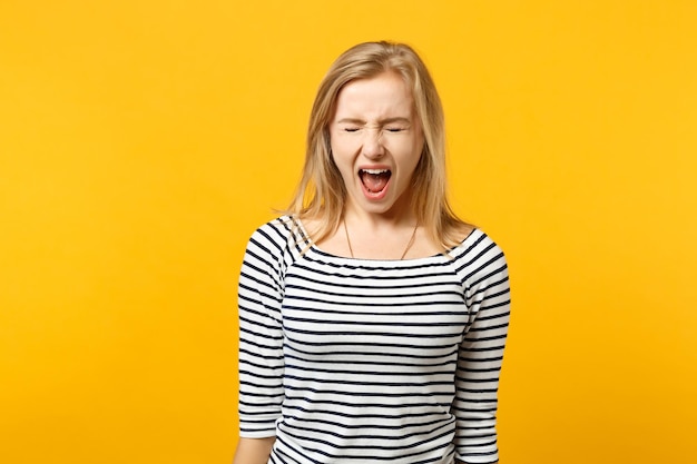 Portrait of crazy young woman in striped clothes keeping eyes closed and screaming isolated on yellow orange wall background in studio. People sincere emotions, lifestyle concept. Mock up copy space.