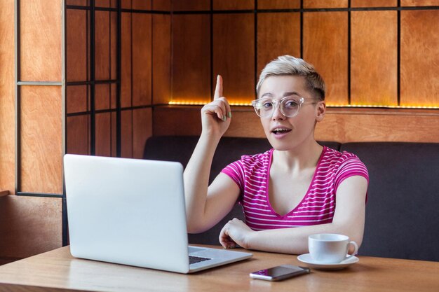 Portrait of creative positive attractive young girl freelancer in pink t-shirt is sitting in cafe and working on laptop with toothy smile and showing finger up, looking at camera. indoor