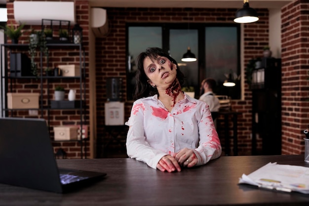 사무실 책상에 앉아 상처와 흉터로 끔찍하게 보이는 소름 끼치는 여성 좀비의 초상화. 무서운 잔인한 죽은 시체가 뇌를 먹고 공격적이고 불길한 피투성이의 악몽을 꾸고 있습니다.