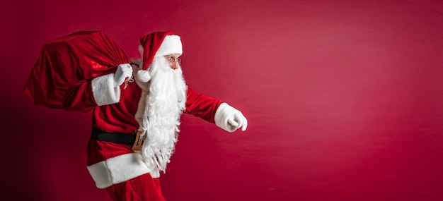 Portrait of crouching Santa Claus carrying huge red sack with presents on red studio background. Isolate