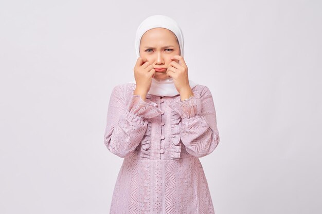 Portrait of crying beautiful young Asian Muslim woman wearing hijab and purple dress rubbing tears wants to cry depressed and has a problem isolated on white studio background