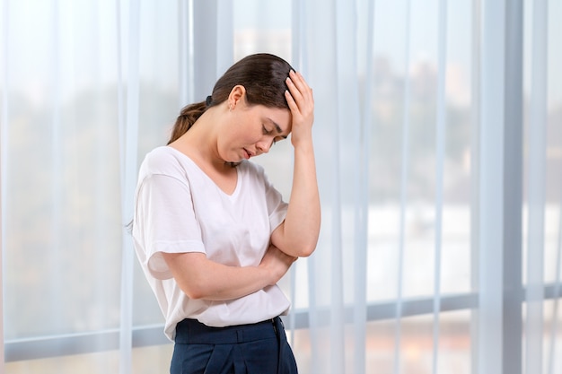 portrait of crying woman holding her head with her hand. in the background of the window. copy space. the concept of domestic violence.